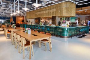 Modern café with wooden tables and chairs, a green-tiled bar with high stools, hanging gold mesh decor, floral arrangements on tables, and bright overhead lighting in a spacious, stylish interior.