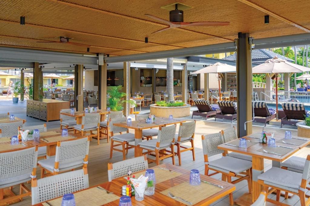 An open-air restaurant with wooden tables and chairs set for dining, overlooking a pool area with lounge chairs and umbrellas, surrounded by greenery and a relaxed, sunny atmosphere.
