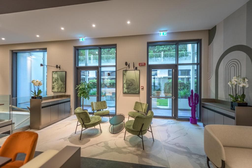 Modern lobby with green chairs and round tables, large windows, wall art, potted plants, and a purple decorative cactus. Soft lighting and neutral tones create a welcoming atmosphere.