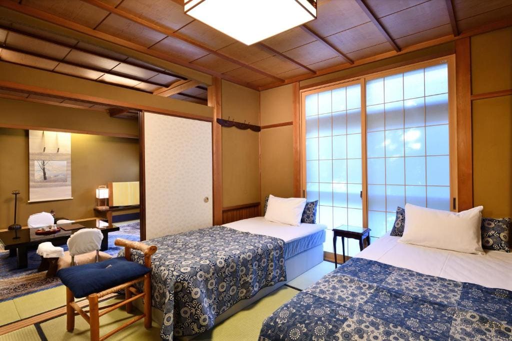 A traditional Japanese-style room with two single beds, blue patterned bedding, tatami mat flooring, shoji paper sliding doors, and a low table with floor cushions in an adjoining space.