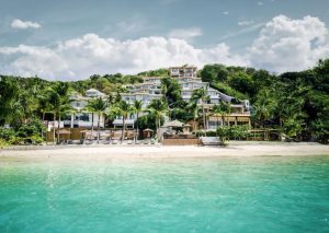 A luxurious beachfront resort with multiple white buildings and palm trees sits on a sandy beach, overlooking clear turquoise water under a partly cloudy sky.