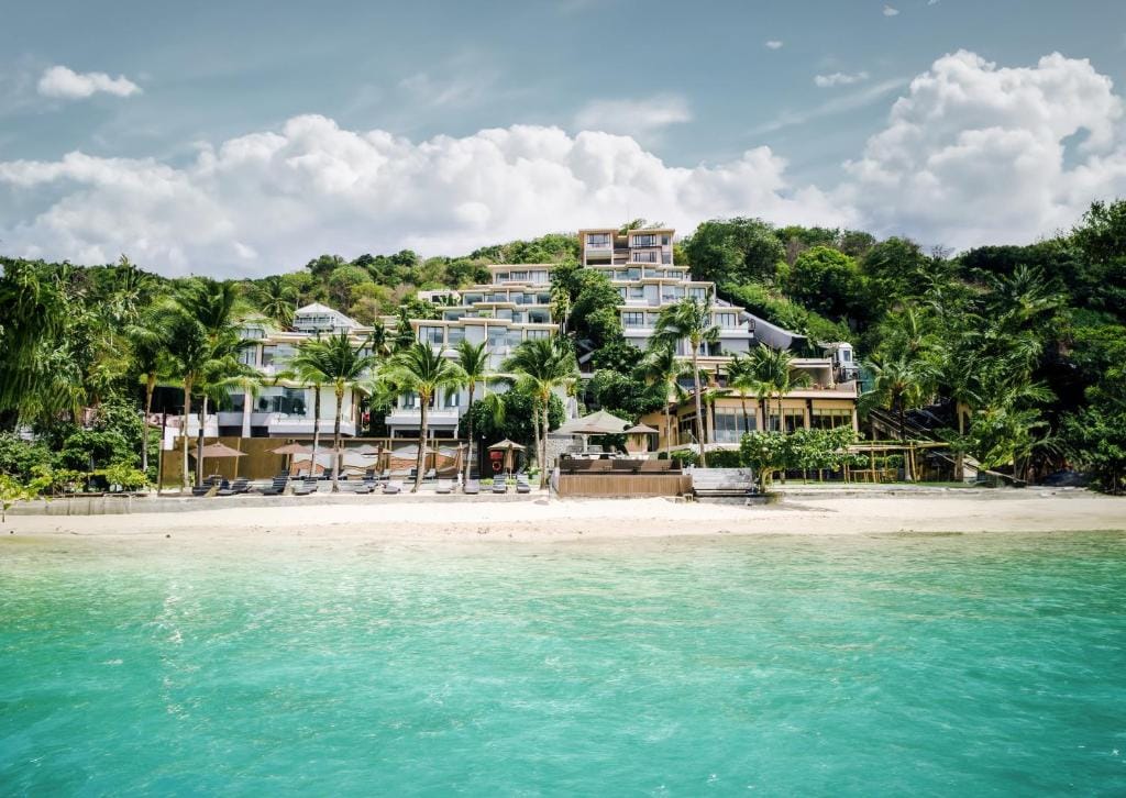 A luxurious beachfront resort with multiple white buildings and palm trees sits on a sandy beach, overlooking clear turquoise water under a partly cloudy sky.