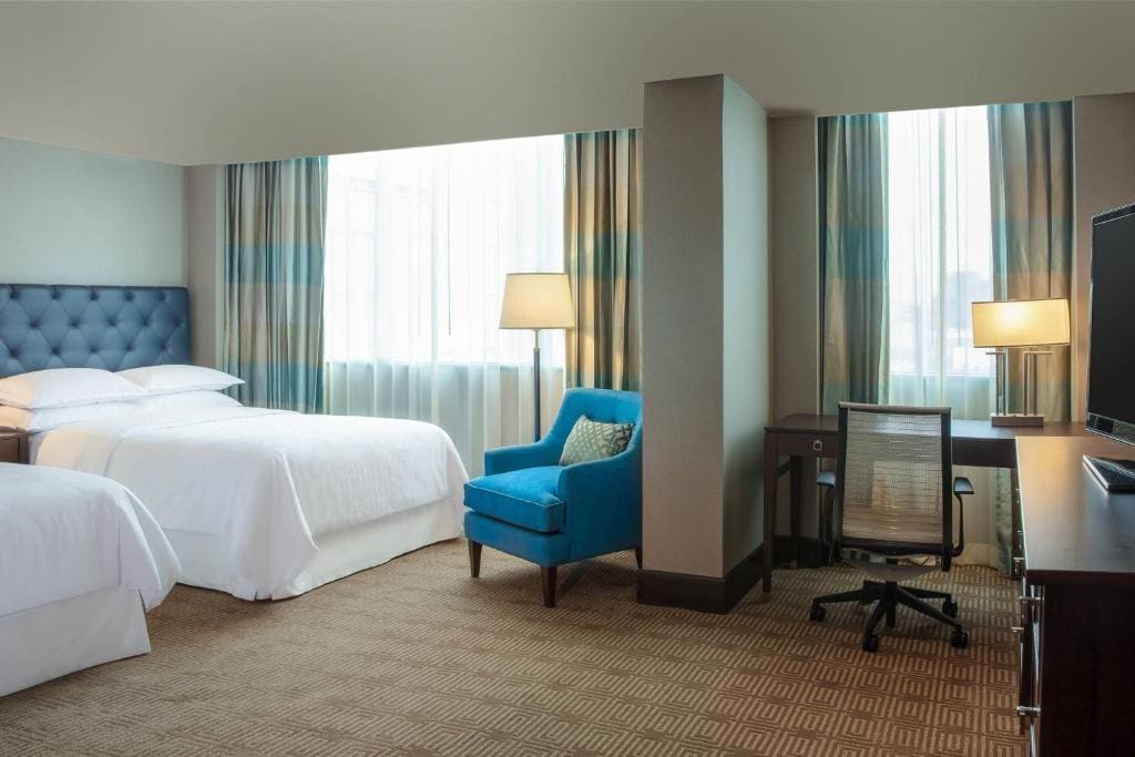 Hotel room with two white beds, a blue armchair, a desk with a chair and lamp, and large windows with striped curtains letting in natural light. A TV sits on a dark wood dresser.