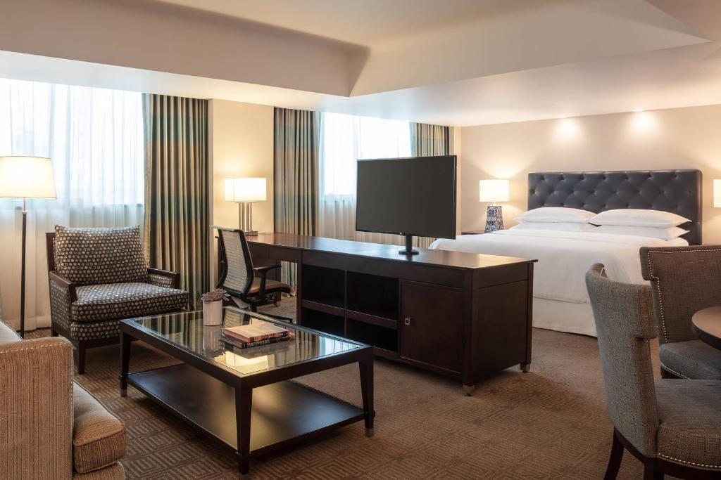 A spacious hotel room with a king bed, modern armchairs, a glass coffee table, a work desk with a chair, and a TV on a stand, featuring large windows with striped curtains and neutral-toned decor.