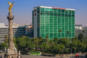 A large green Sheraton Maria Isabel hotel building stands behind trees, with the golden Angel of Independence monument in the foreground in Mexico City.