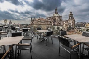 Outdoor terrace with empty tables and chairs, overlooking a historic cathedral under a dramatic, cloudy sky, with city buildings visible in the distance.