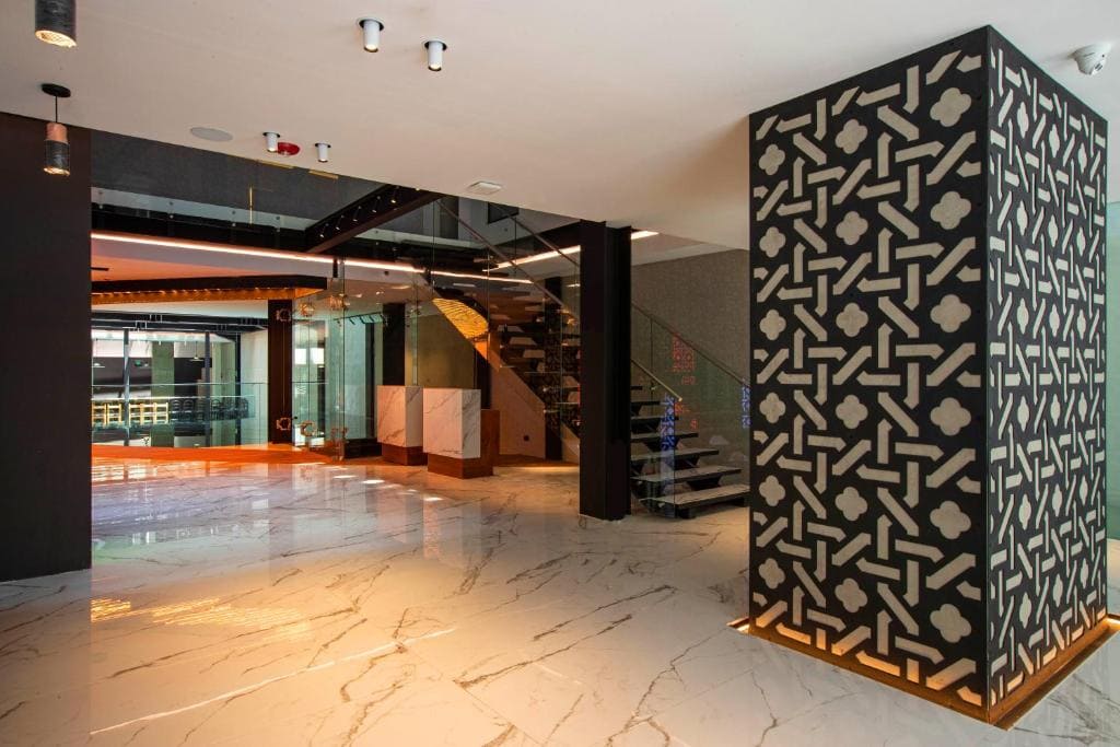 A modern, spacious lobby with glossy marble floors, a decorative patterned pillar, glass railings, and a staircase leading to an upper level. Warm lighting and sleek design elements highlight the contemporary style.
