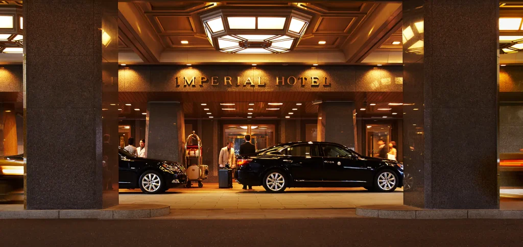 Two black luxury cars are parked under the covered entrance of the Imperial Hotel at night, with hotel staff and guests near the door and warm lighting illuminating the scene.