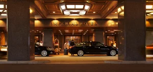 Two black luxury cars are parked under the covered entrance of the Imperial Hotel at night, with hotel staff and guests near the door and warm lighting illuminating the scene.