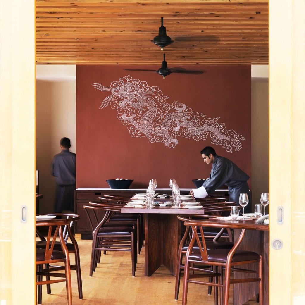 A server sets a table in a stylish restaurant with wooden chairs and tables. The back wall features a large white dragon mural on a reddish-brown background. Another person stands near the wall.