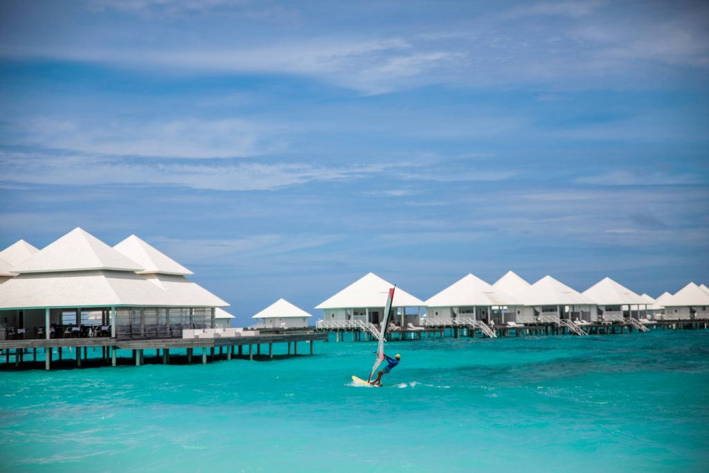 A person windsurfing on bright turquoise water near white overwater bungalows under a blue sky with scattered clouds.