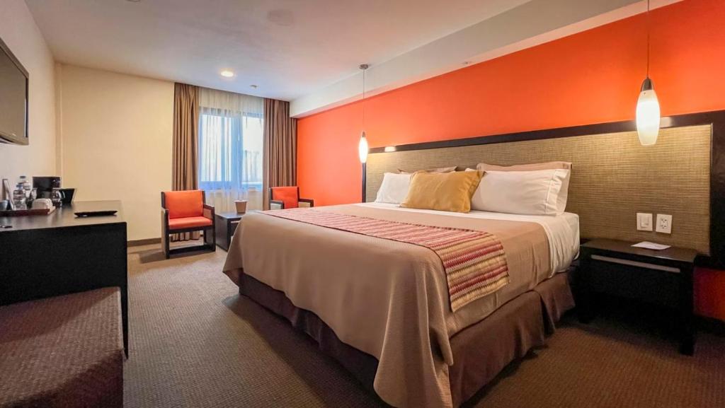 A hotel room with a king-size bed, orange accent wall, bedside lamps, two chairs and a small table by the window, a desk with a TV, and neutral-colored carpet.