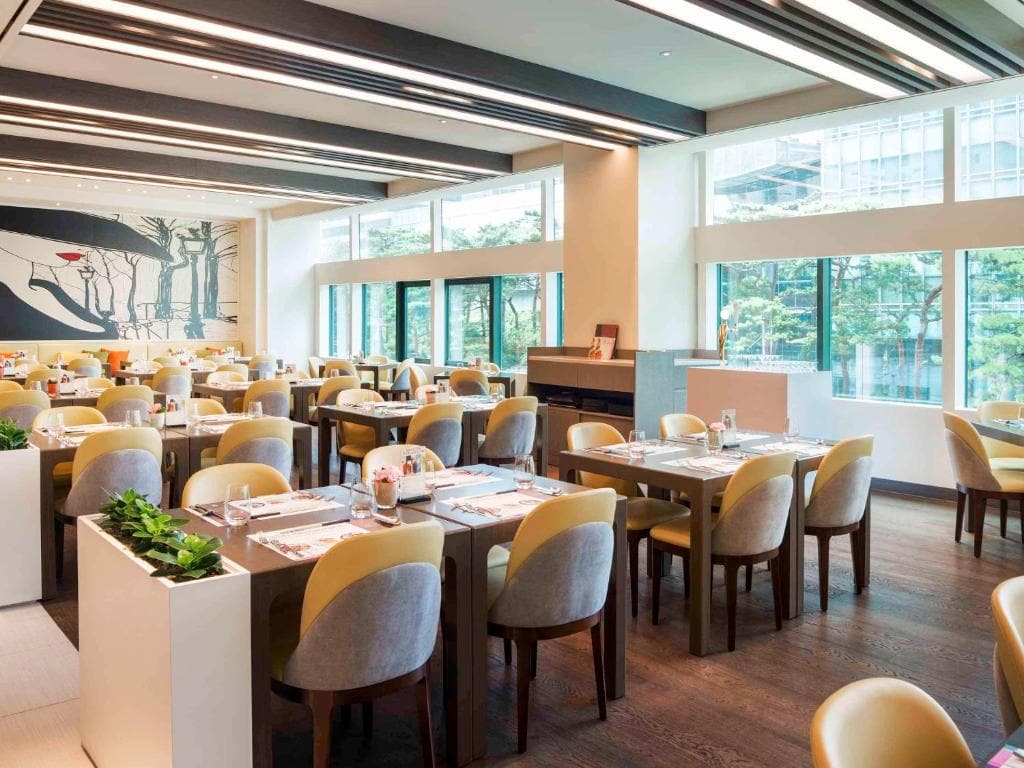 A bright, modern restaurant dining area with neatly arranged tables and chairs, large windows letting in natural light, and wall art depicting abstract tree designs.