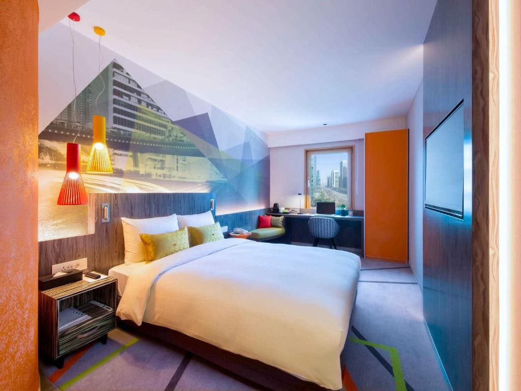 Modern hotel room with a large bed, colorful pillows, unique wall art, hanging lamps, a desk by the window, and city buildings visible outside. The room has a vibrant and contemporary design.