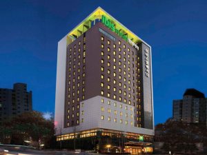 A tall, modern hotel building illuminated at night, with many lit windows and signage in Korean on the facade. The hotel is surrounded by trees and other buildings, with a deep blue evening sky overhead.