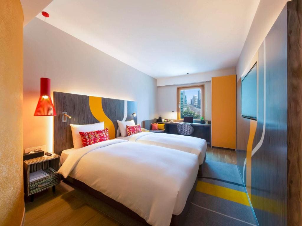 Modern hotel room with two single beds, vibrant pillows, a red hanging lamp, a large window with a city view, a wall-mounted TV, a desk, and colorful decor accents in yellow and gray tones.