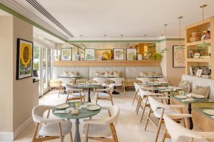 A bright, modern café with light-colored walls, green accents, round tables, and white chairs. Artworks and plants decorate the shelves and walls, while large windows let in natural light.