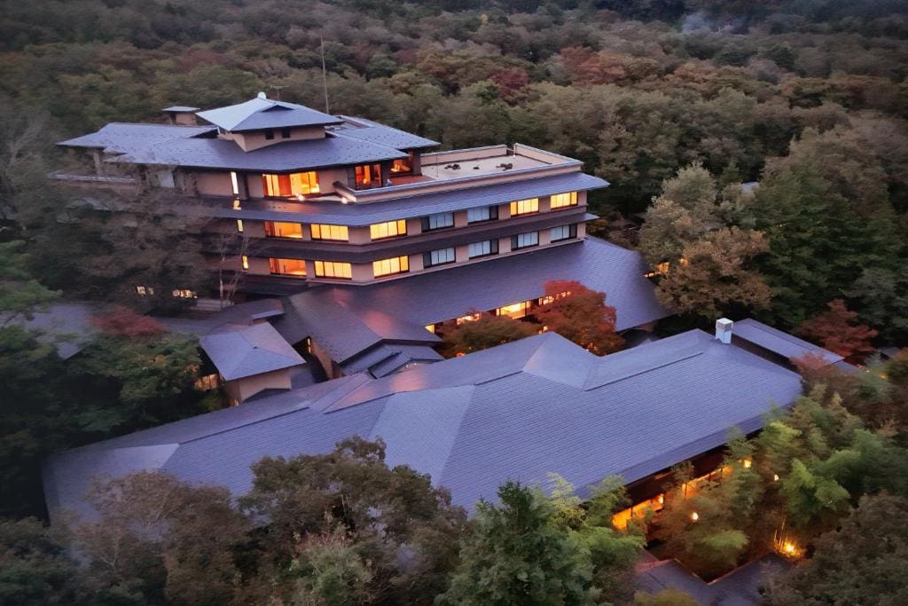 A multi-story building with traditional Japanese architectural design, surrounded by dense trees, is illuminated warmly from within at dusk. The rooftop features layered, angular roofs and scenic natural surroundings.
