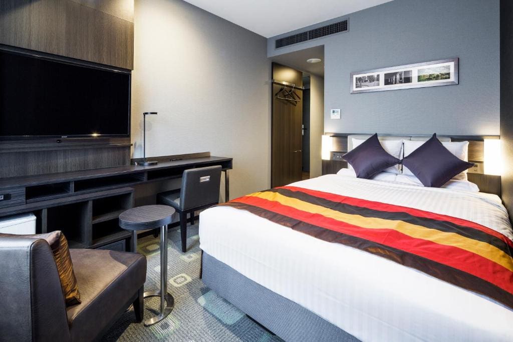 Modern hotel room with a double bed, colorful striped runner, two dark pillows, a desk with chair, large TV, small round table, armchair, and a closet area in the back under a framed photo.