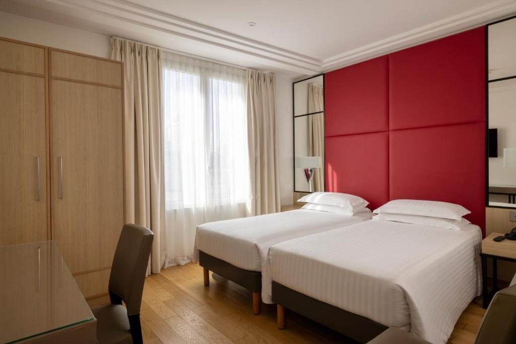 A modern hotel room with two single beds, white bedding, a large red padded headboard, light wood furniture, a desk with a chair, and a window with beige curtains letting in natural light.