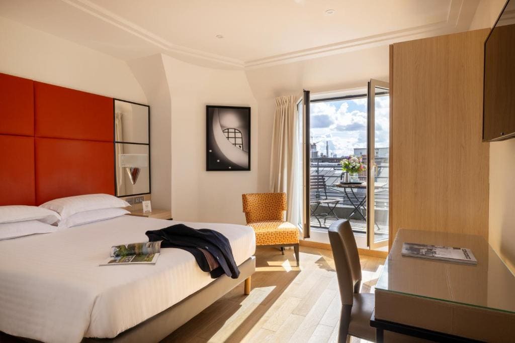 A bright hotel room with a large bed, desk, chair, and TV. Glass doors open to a balcony with a table, two chairs, and city views under a partly cloudy sky. A magazine and jacket are on the bed.