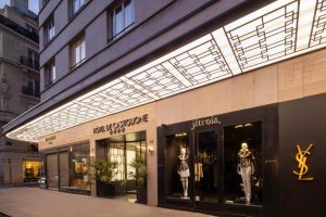 A modern street view of upscale storefronts, including Saint Laurent and Jitrois, with mannequins in stylish clothing and a bright, geometric overhang above the entrances.