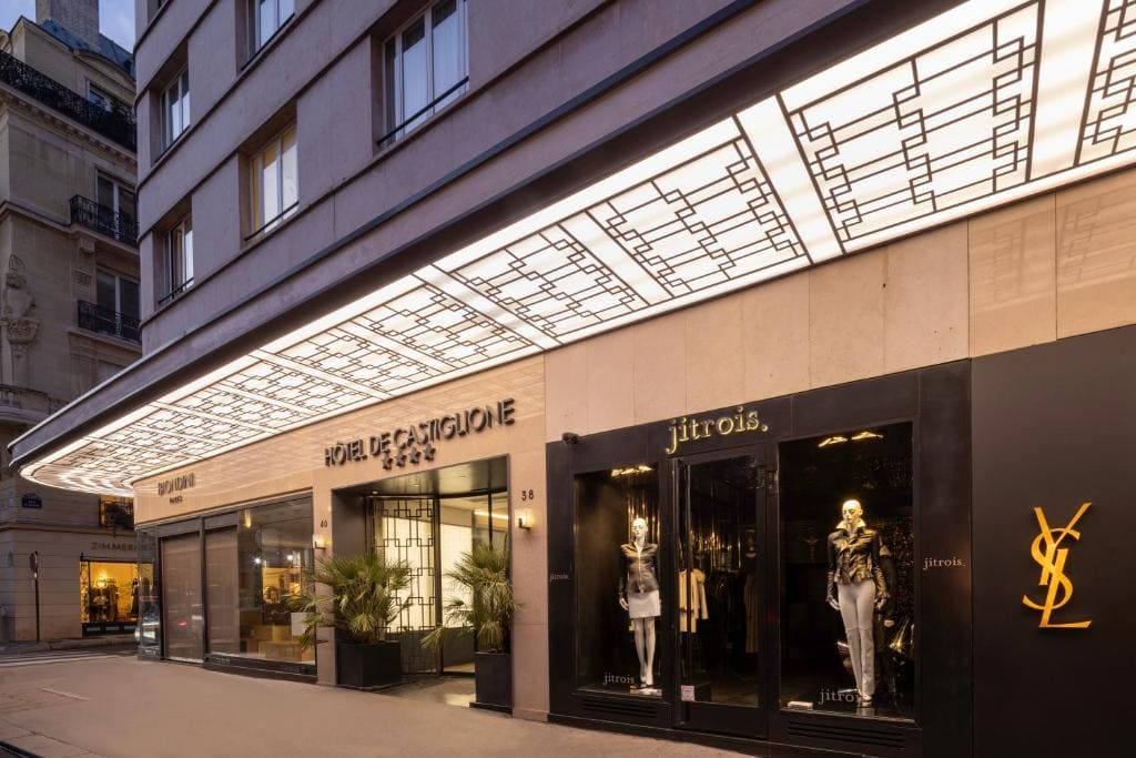 A modern street view of upscale storefronts, including Saint Laurent and Jitrois, with mannequins in stylish clothing and a bright, geometric overhang above the entrances.