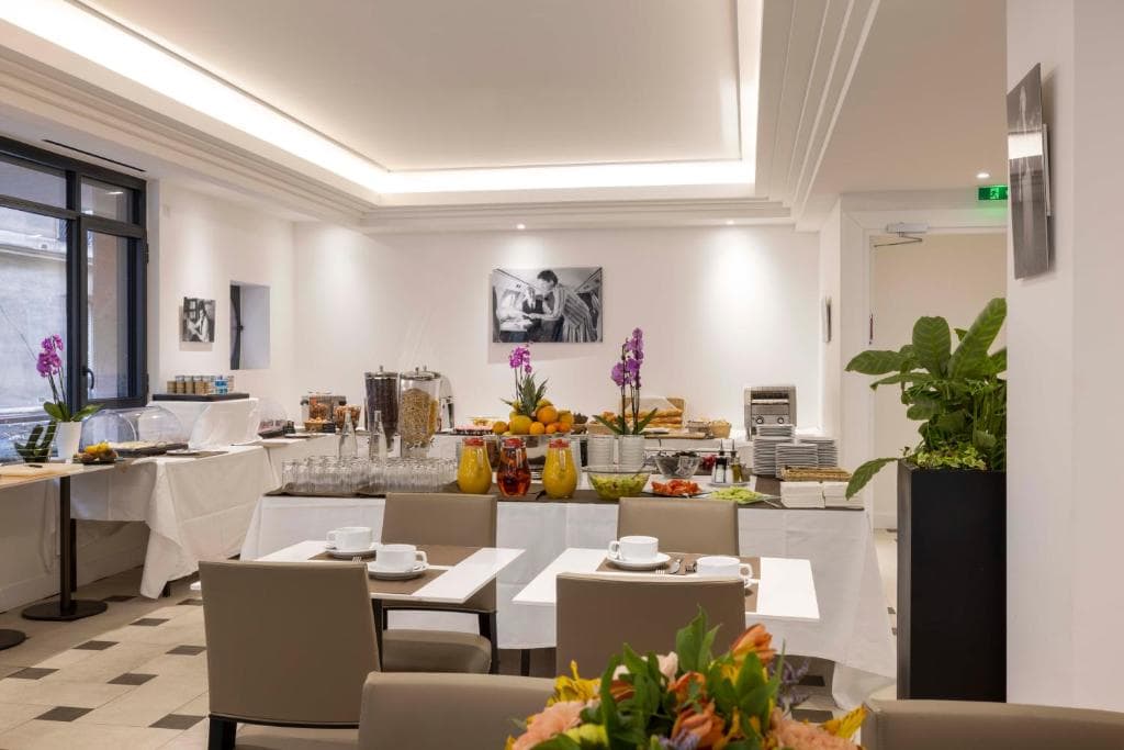 A bright, modern breakfast buffet room with tables set for dining, a buffet table with cereals, fruit, juices, pastries, and plates, decorated with orchids and green plants. Black-and-white photos hang on the white walls.