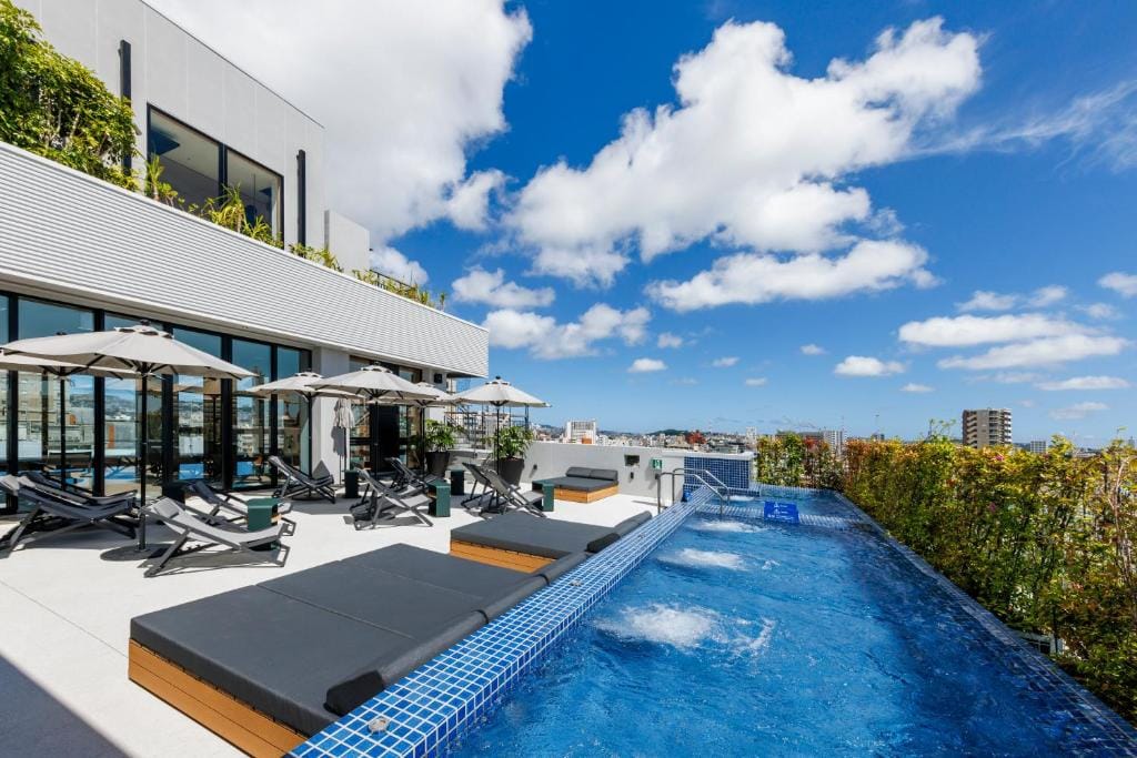 A rooftop pool with blue tiles has lounge chairs and umbrellas along the side, next to a modern building with large windows. Greenery surrounds the area under a bright blue sky with scattered clouds.