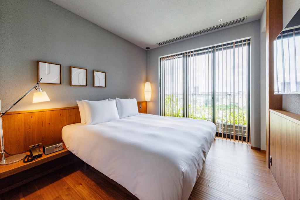 A modern hotel room with a large bed, white bedding, wooden floors, and minimal decor. There are three framed pictures on the wall, a bedside lamp, and large windows with vertical blinds letting in natural light.