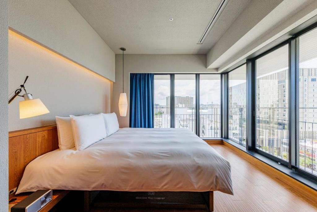 A modern hotel room with a large bed, white bedding, a wooden headboard, a wall lamp, and floor-to-ceiling windows offering a city view. Blue curtains are partially drawn to the side.