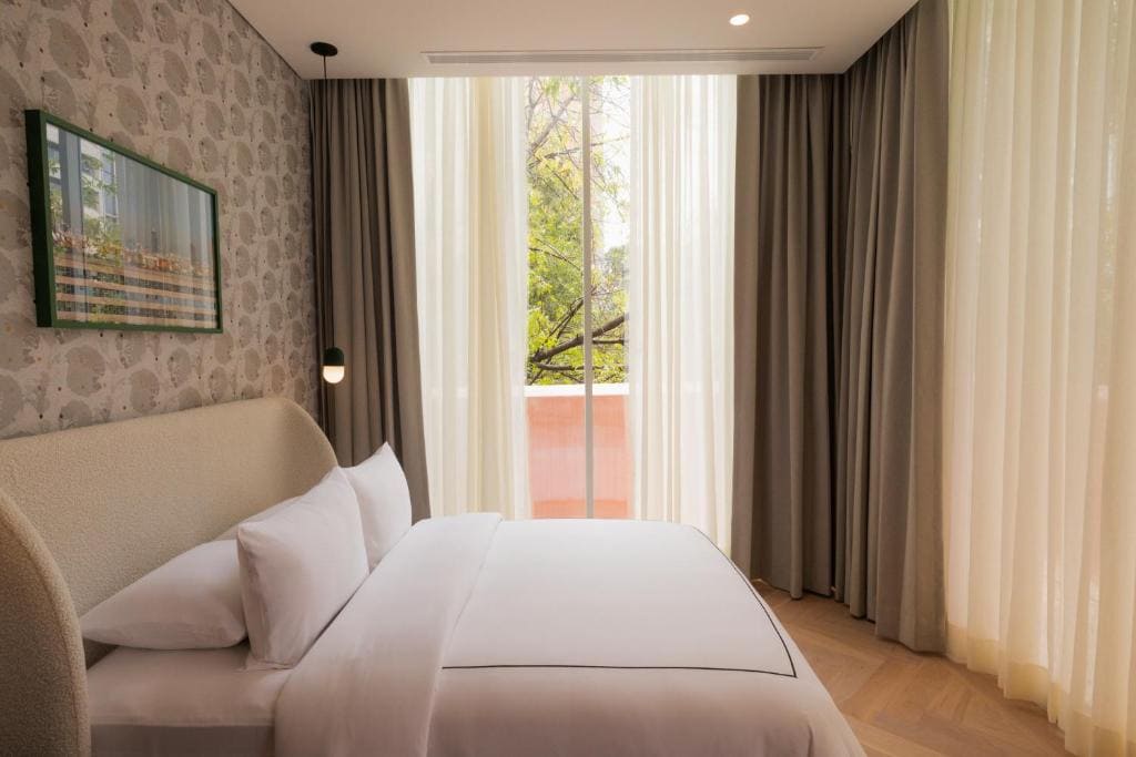 A cozy bedroom with a double bed, white bedding, patterned wallpaper, a framed picture, and large floor-to-ceiling windows with sheer curtains letting in natural light and a view of green trees outside.