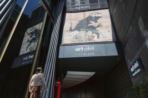 A person looks up at a large mural of a rat with a guitar, painted above the entrance of the art’otel London Hoxton. The building’s modern facade and hotel signage are visible.