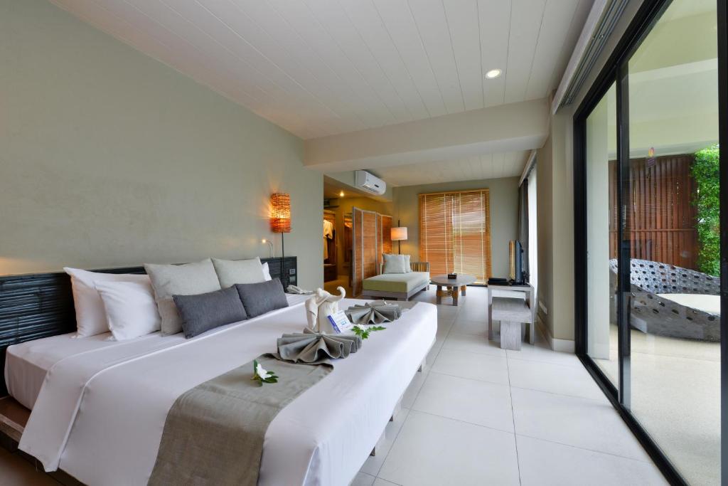A modern hotel room with a large bed, neutral decor, two towel swans, a couch, TV, and floor-to-ceiling windows leading to a private outdoor seating area. Light fills the room through the glass doors.