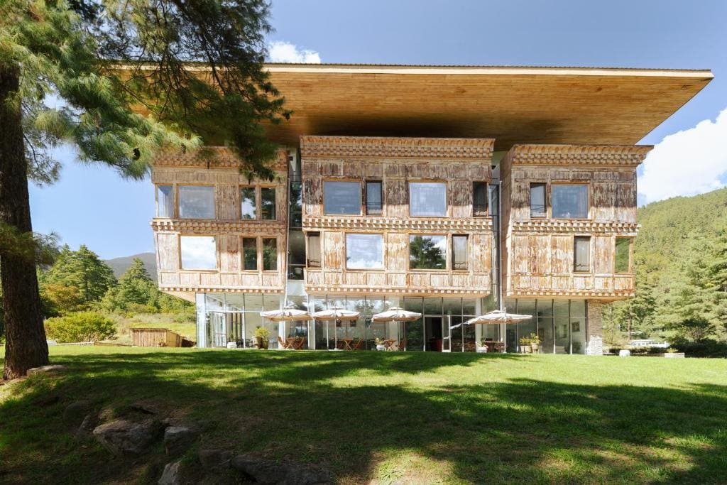 A large building with ornate wooden facades, large windows, and a wide flat roof, sits on a grassy lawn with outdoor tables and umbrellas, surrounded by trees and mountains.