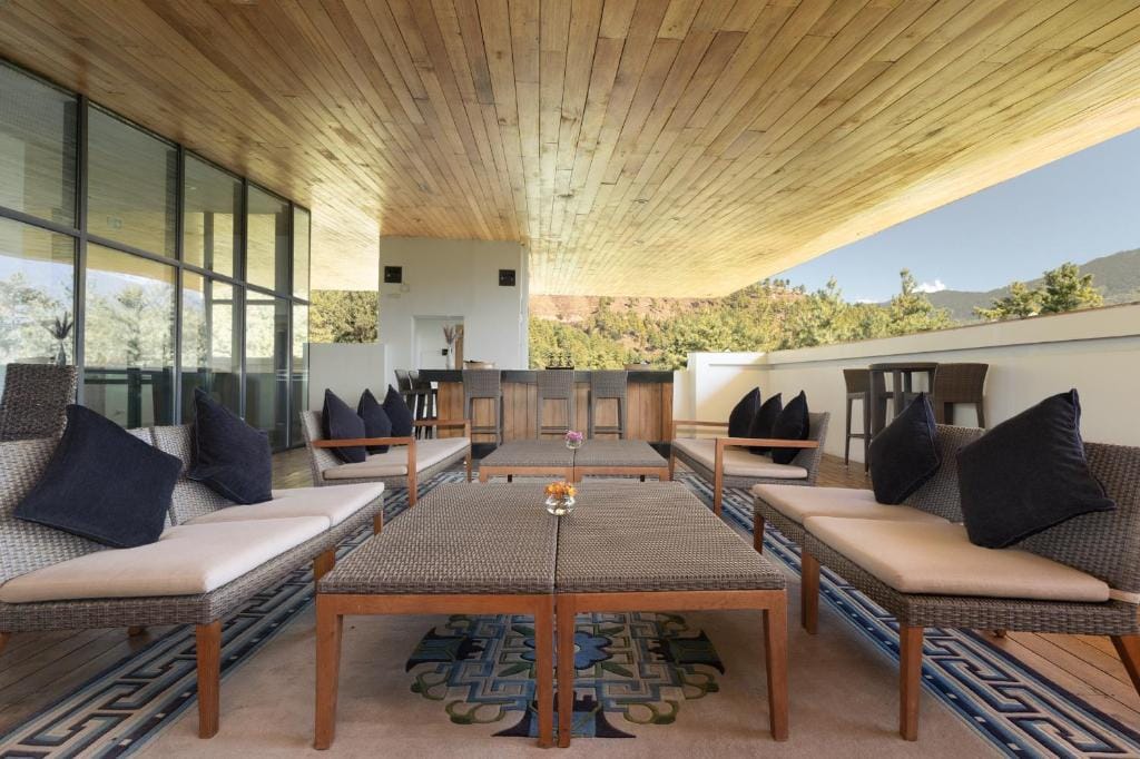 Modern outdoor lounge with wicker furniture, cushioned seats, dark throw pillows, low tables, and a patterned rug. The space has a wooden ceiling and open views of trees and mountains in the background.
