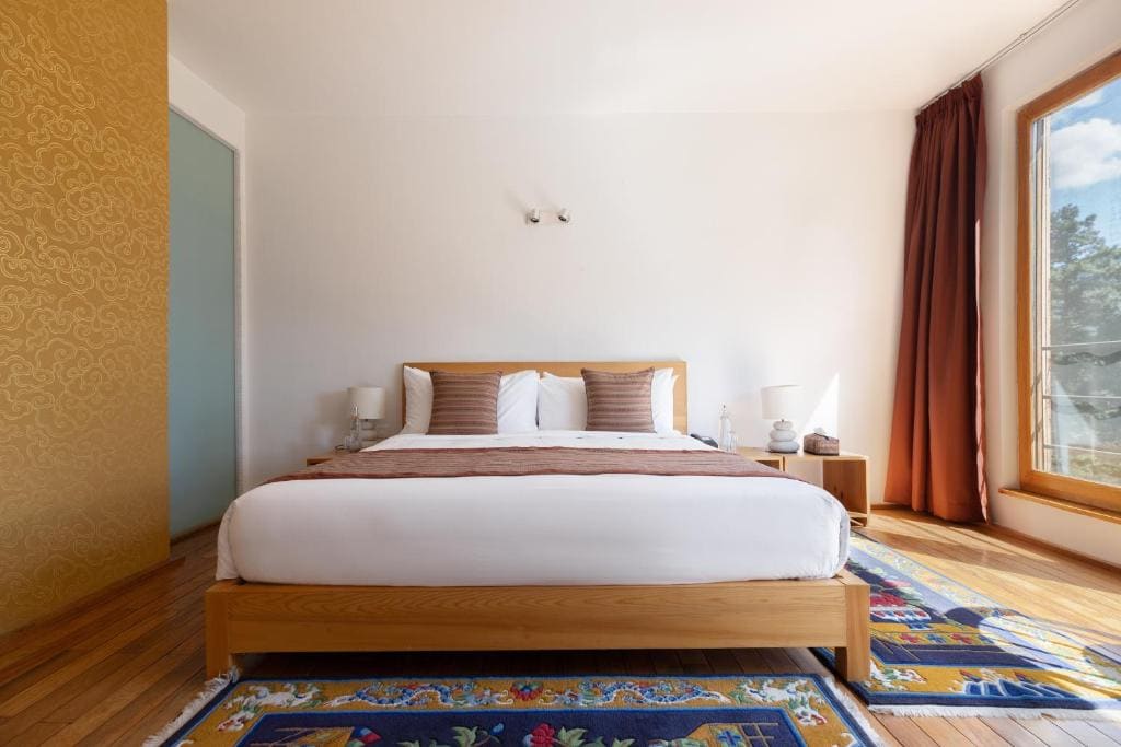 A bright, modern bedroom with a wooden bed, white bedding, and brown accent pillows. Patterned colorful rug, bedside tables with lamps, large window with brown curtains, and sunlight streaming in.