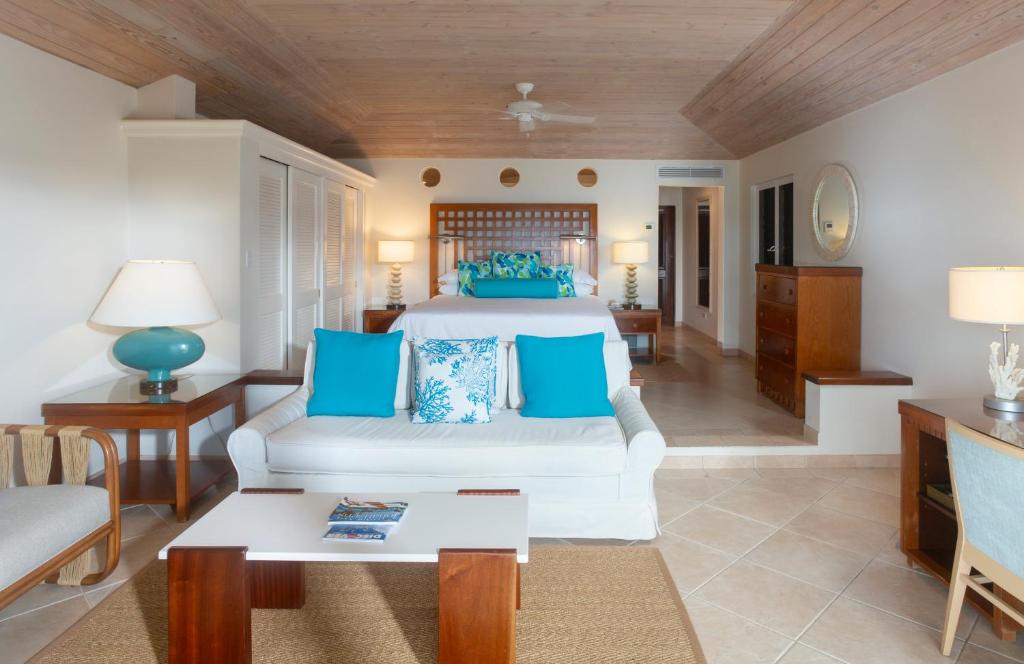Spacious, airy bedroom with a bed featuring blue and white pillows, a white sofa, wooden furniture, lamps, a ceiling fan, and light tile flooring under a wooden ceiling. The room has a cozy, beachy decor.