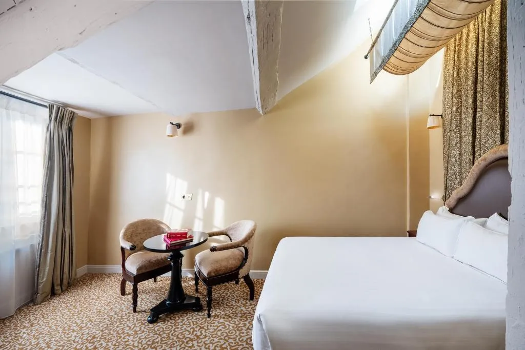 A cozy hotel room with a neatly made bed, two upholstered chairs around a small round table, beige walls, patterned carpet, and sunlight streaming in through a window with light curtains.
