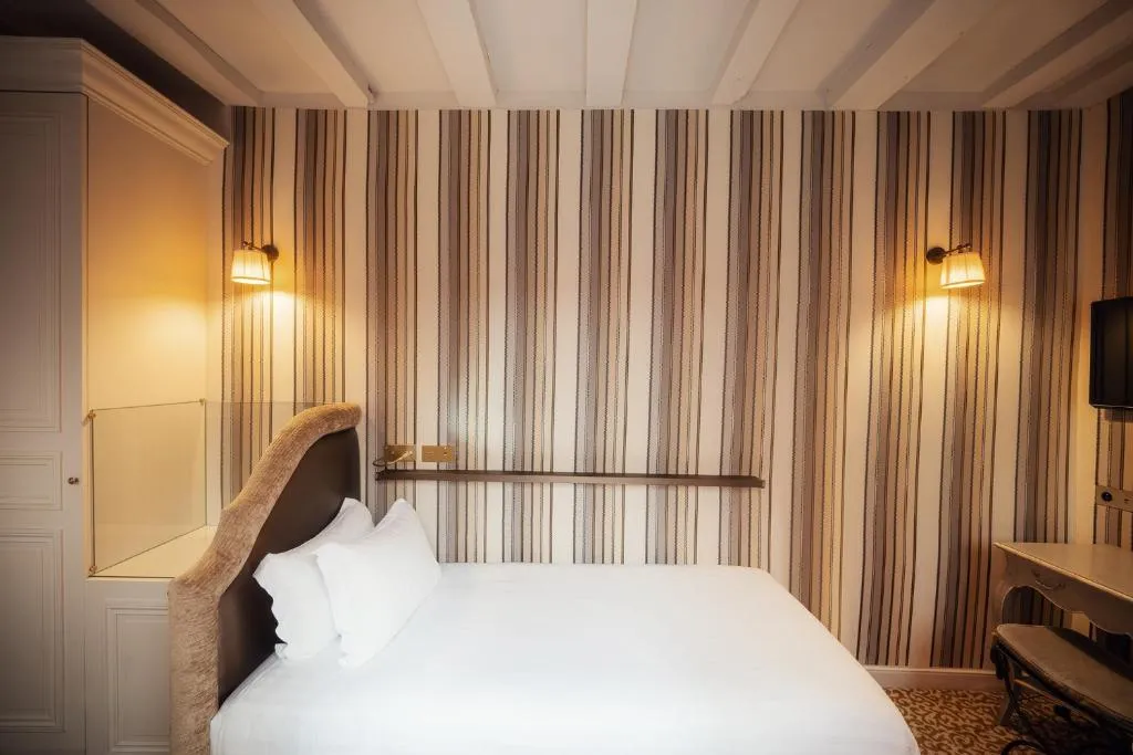 A small, neatly made bed with white linens sits in a cozy room with striped wallpaper, two wall lamps, a mounted TV, and a built-in shelf above the bed’s headboard.