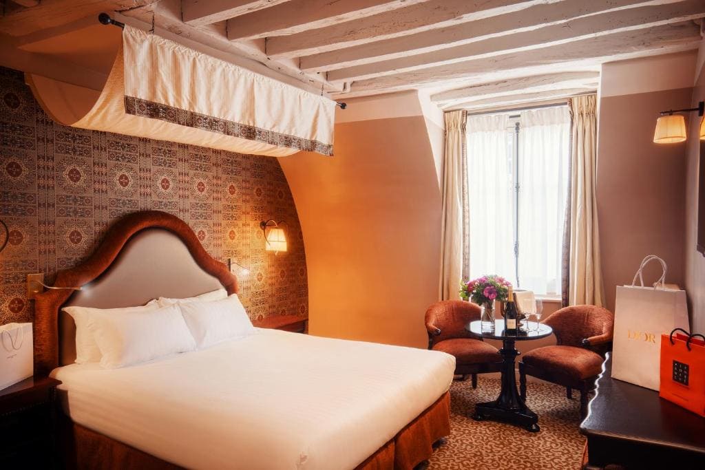 A cozy hotel room with a large bed, patterned wallpaper, two armchairs and a small round table with flowers, a window with curtains, and shopping bags on the side table. Exposed ceiling beams add charm.