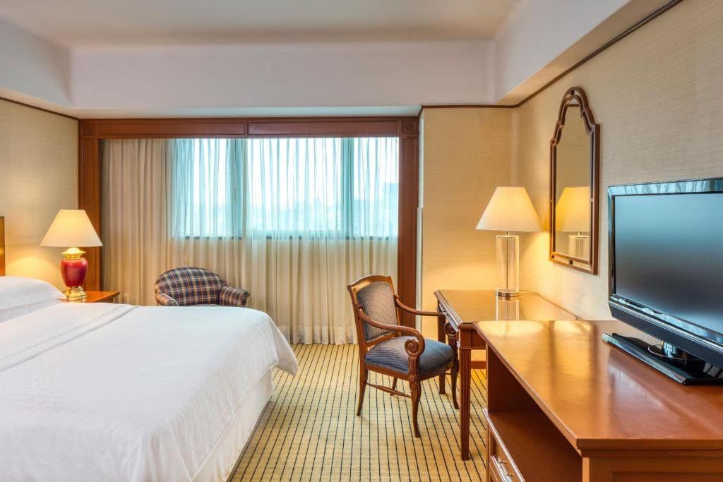 A hotel room with a large bed, desk and chair, flat-screen TV, two lamps, an armchair by the window, and light-colored curtains letting in natural light.
