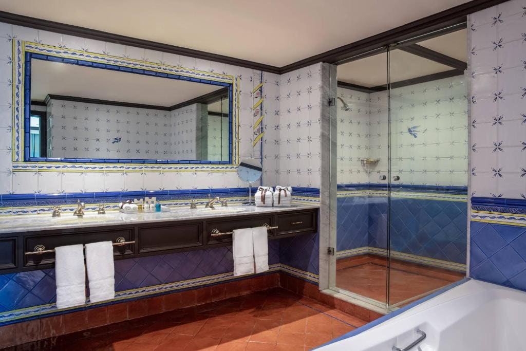 Elegant bathroom with a large mirror, double sinks, and a glass shower. The walls are decorated with blue and white tiles featuring a star pattern, and towels hang neatly under the sink. Part of a bathtub is visible.