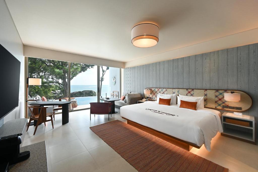 Spacious modern hotel room with a large bed, seating area, desk, and floor-to-ceiling windows offering a view of trees and the ocean. The décor is minimalistic with warm tones and contemporary furnishings.
