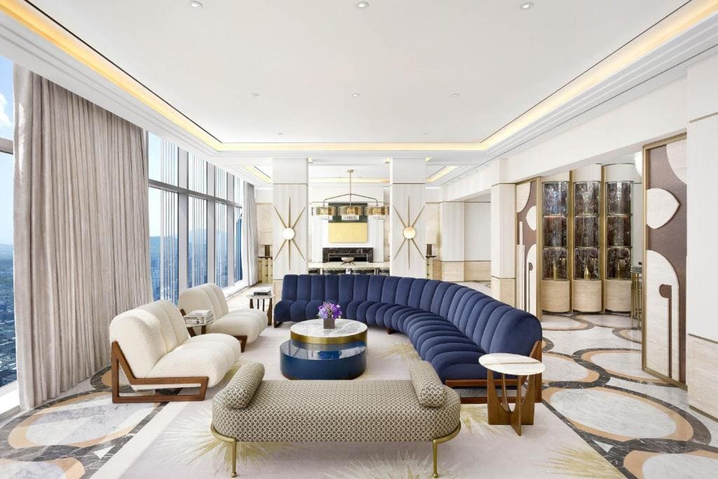 Spacious, modern living room with large windows, curved blue sofa, white armchairs, patterned rug, round coffee table, and elegant light fixtures; city view visible through floor-to-ceiling windows.