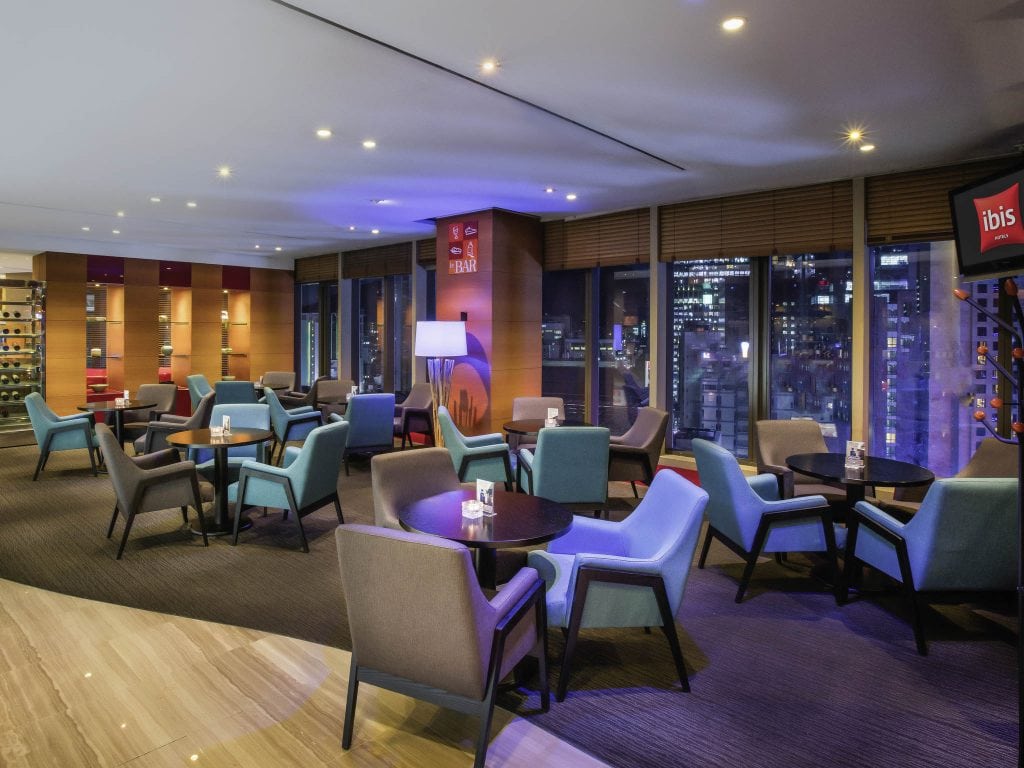 Modern hotel lounge with blue and gray armchairs, small round tables, floor lamps, and large windows showing a cityscape at night. Wooden shelves and soft lighting create a cozy, inviting atmosphere.