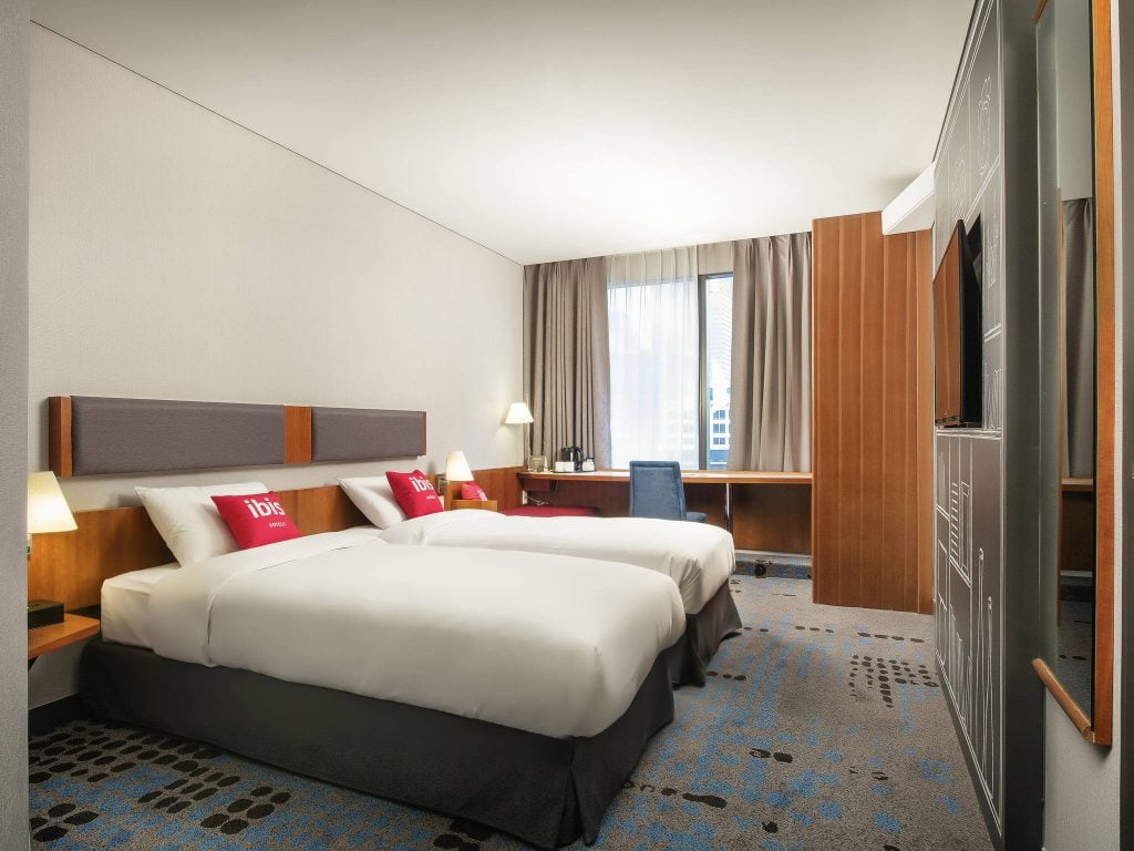 Modern hotel room with two twin beds, red accent pillows, wall-mounted lamps, a desk with a chair near a large window, neutral-colored walls, and a flat-screen TV opposite the beds.