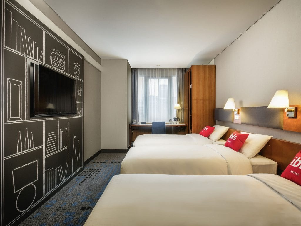 Modern hotel room with two double beds, red accent pillows, a wall-mounted TV, a desk with a chair by the window, and a wall decorated with white line art of books and objects.