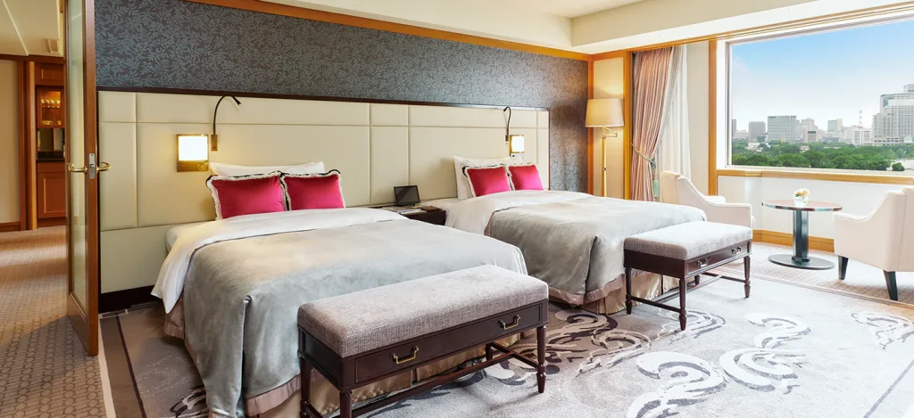 A modern hotel room with two double beds, each with white bedding and red accent pillows. There are benches at the foot of each bed, a large window with a city view, and a seating area with two chairs and a small table.