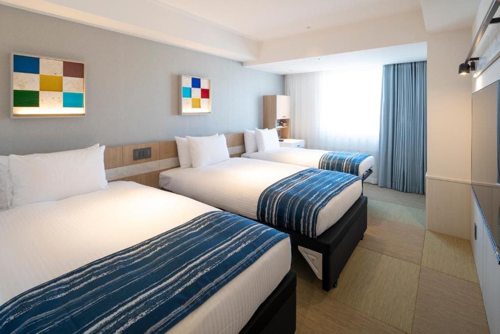 A bright hotel room with three single beds lined up side by side, each with white bedding and blue striped throws. The room has modern decor, a large window with blue curtains, and abstract art on the walls.